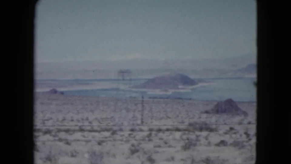 Nevada desert view with sparse vegetation, distant blue water, low mountains, and utility poles or towers visible in the distance.