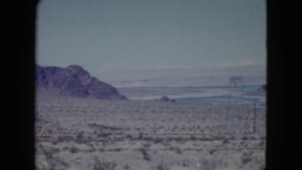 1959 Nevada travel footage frame showing a desert valley, mountain slope, distant water, and utility poles across open terrain.