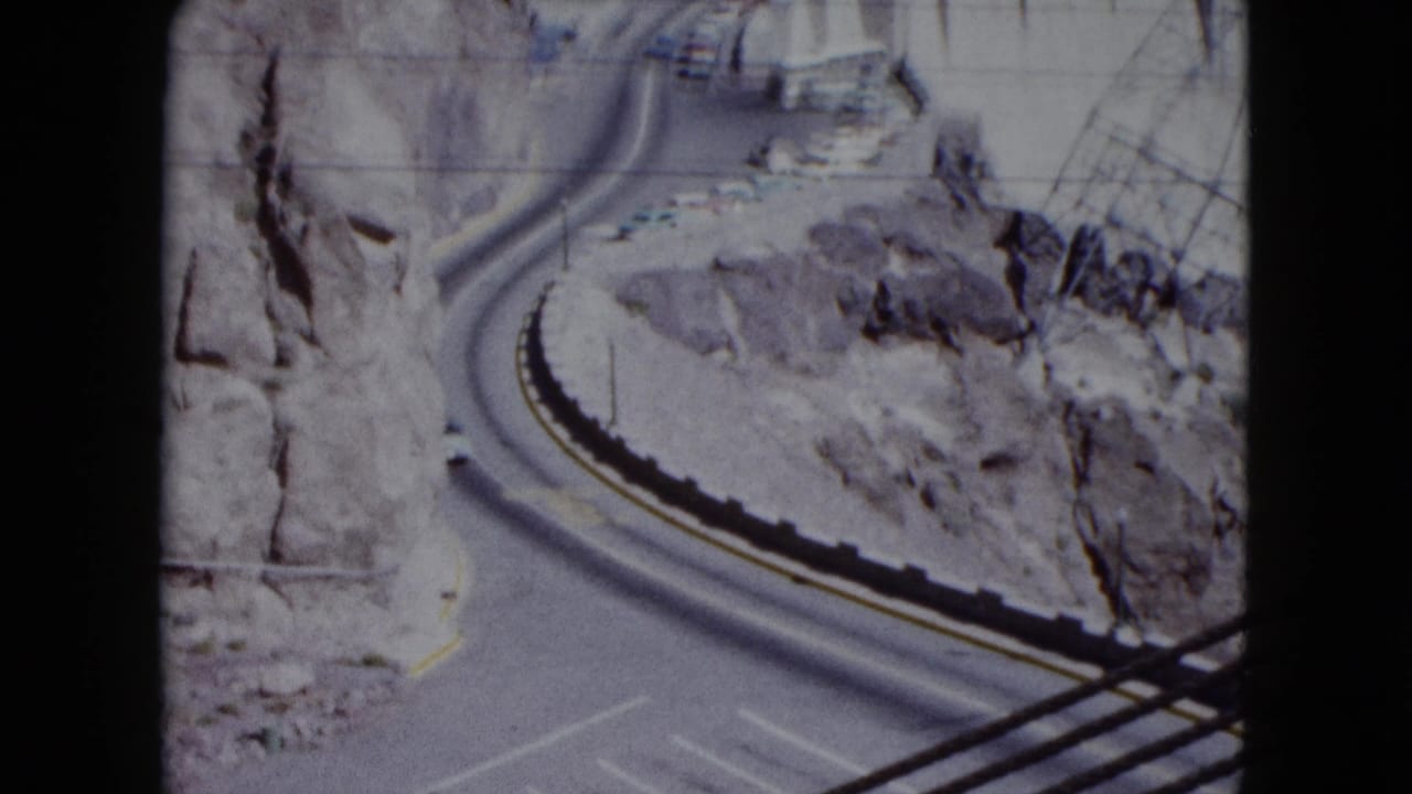 Curving desert road between rocky canyon walls, with part of the dam area beyond.