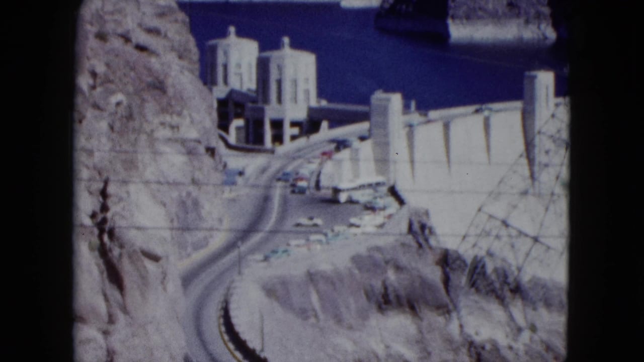 Hoover Dam structures, curving road, cars, rocky cliffs, and blue reservoir water.