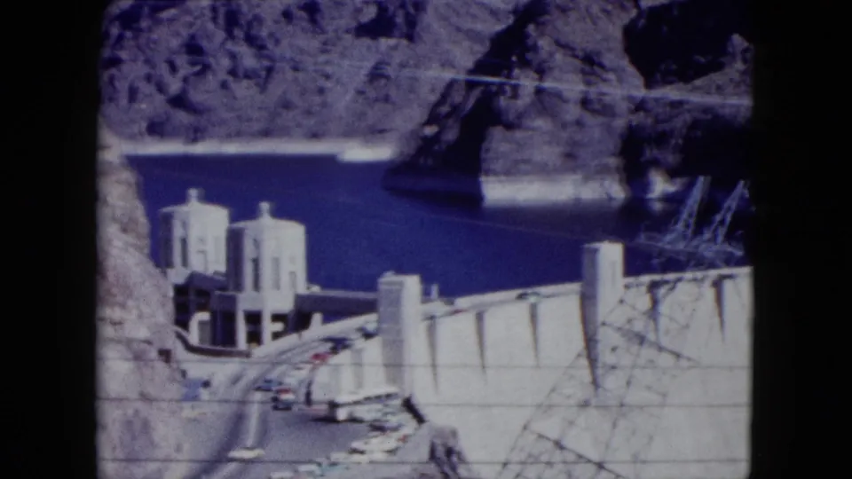 1959 Nevada 8mm frame showing Hoover Dam towers, a road with cars, rocky canyon walls, and blue reservoir water.