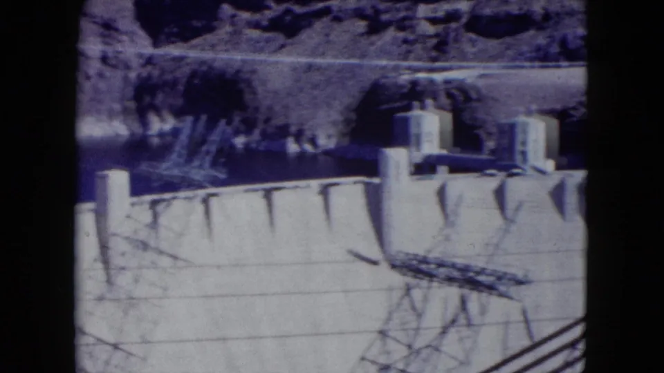 1959 Nevada home-movie frame showing Hoover Dam's concrete wall, intake towers, canyon rim, and dark transmission-line shadows.
