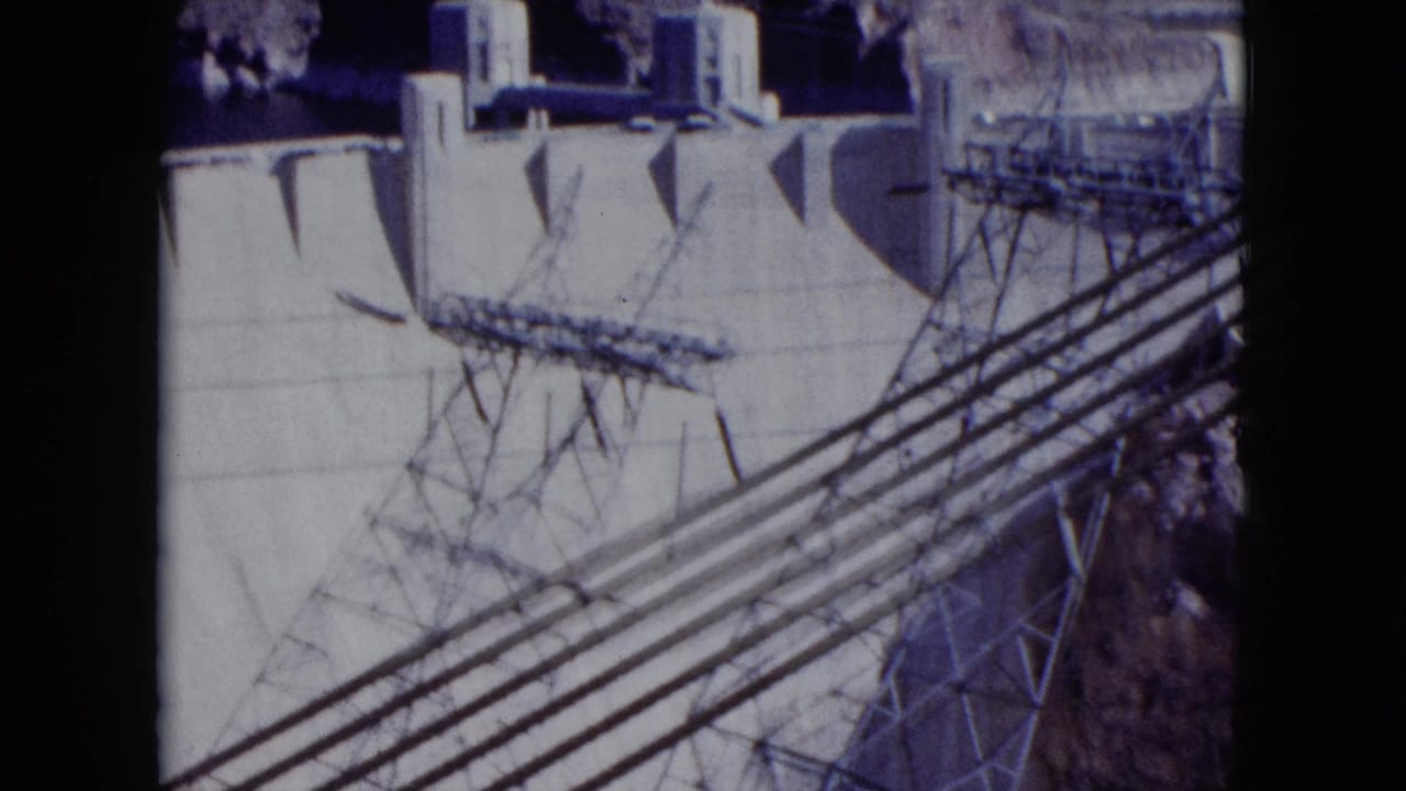 1959 Nevada view of Hoover Dam structures with bundled high-voltage lines and canyon terrain visible around the concrete face.