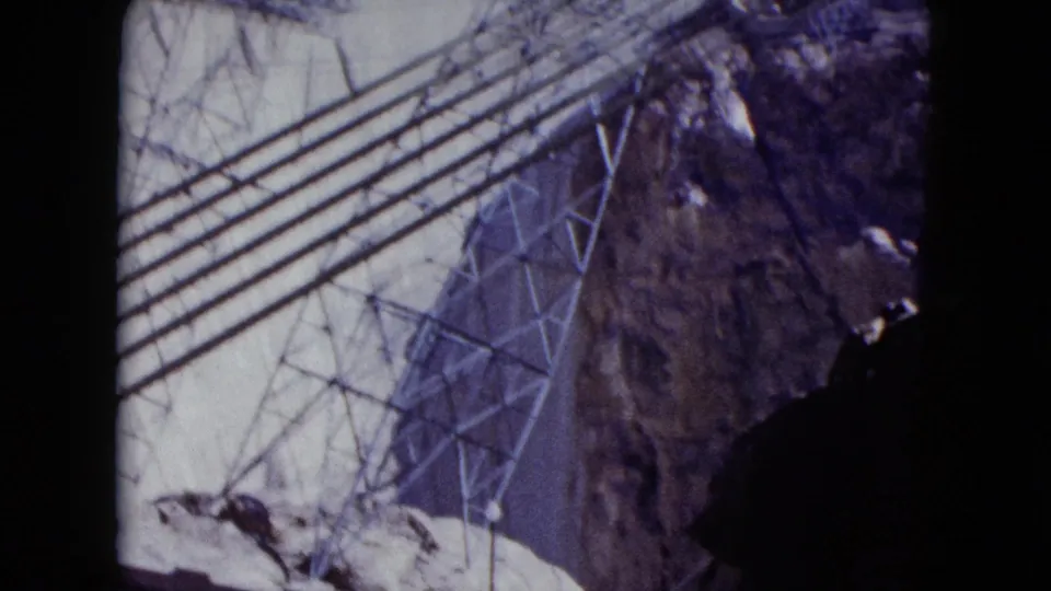 1959 Nevada frame looking across Hoover Dam power infrastructure, with a lattice tower, heavy lines, canyon wall, and empty road.