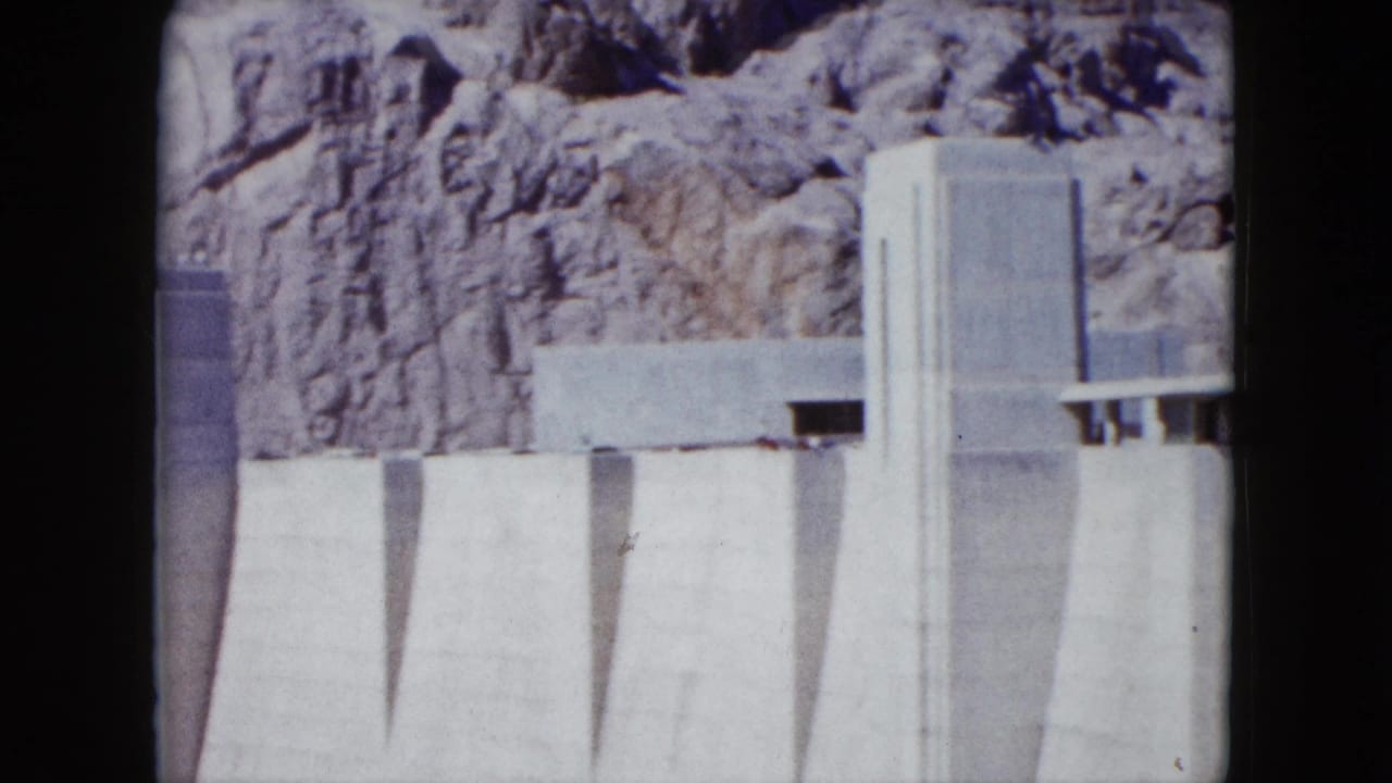 1959 Nevada 8mm frame showing a pale concrete dam face with vertical forms below a rugged canyon wall.