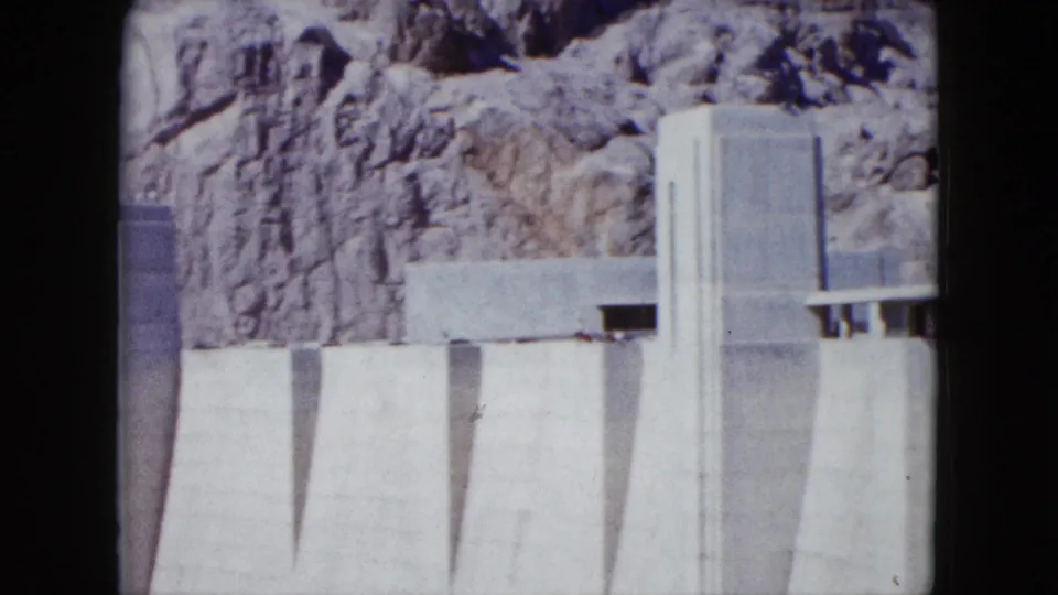 1959 Nevada 8mm frame showing a pale concrete dam face with vertical forms below a rugged canyon wall.