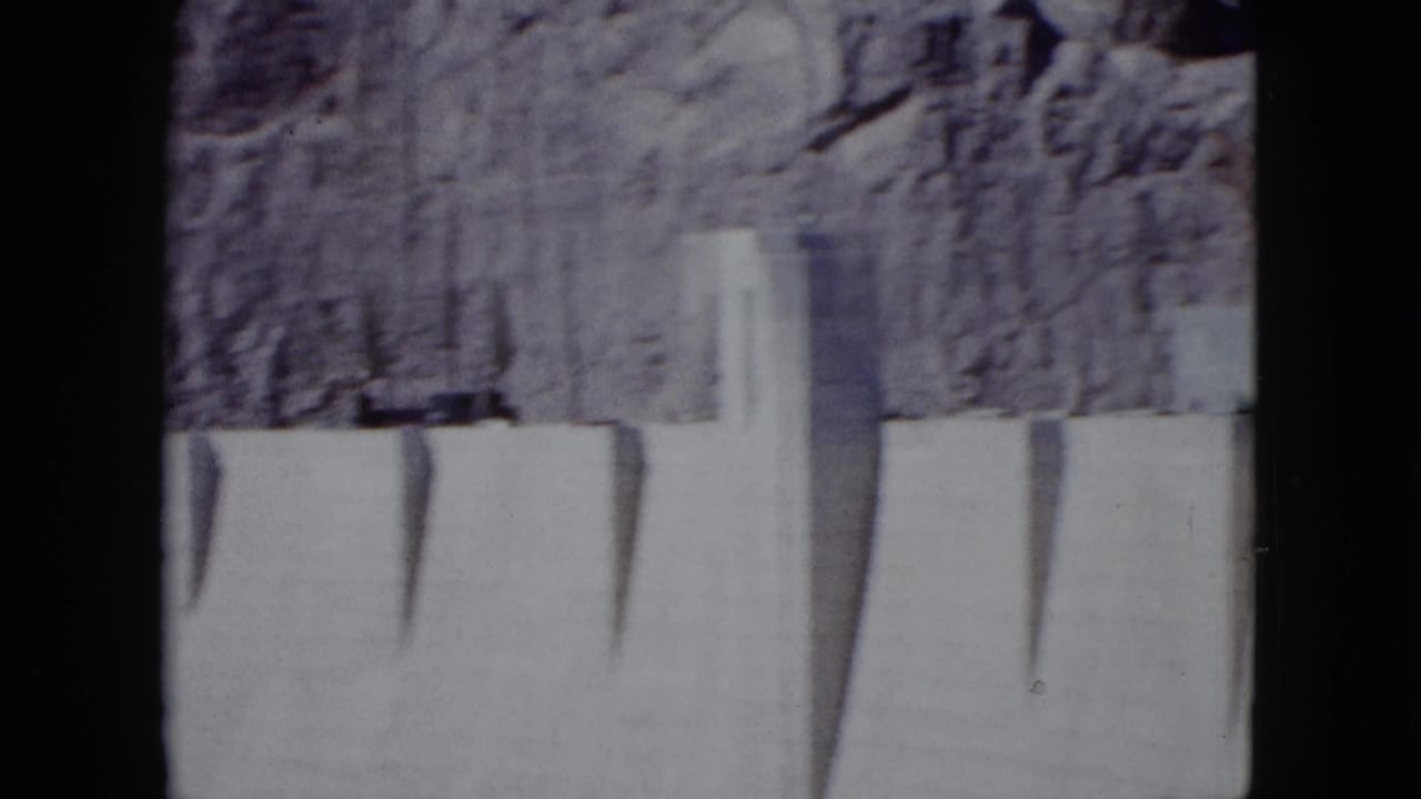 Dam crest and pale concrete wall seen below a rugged rock slope.
