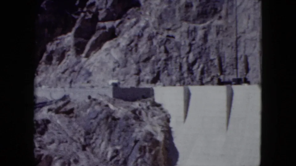 1959 home-movie frame of a concrete dam section and buttress shadows beneath a high Nevada canyon face.