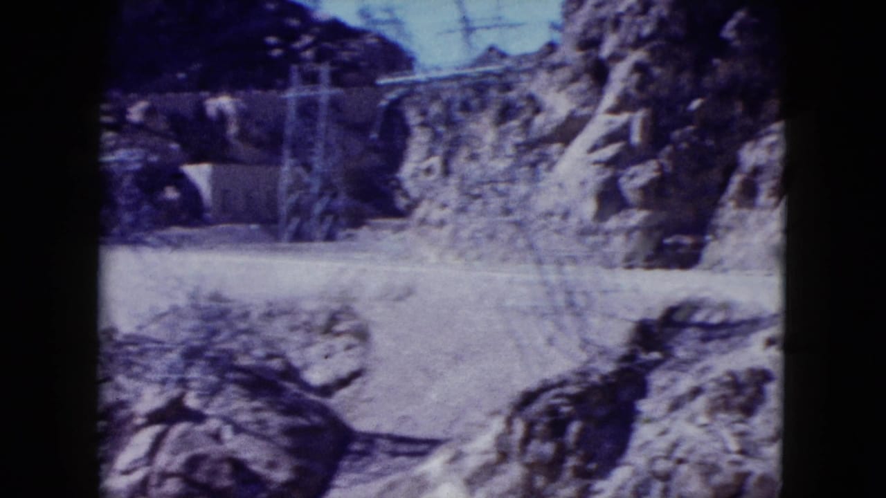 1959 8mm home-movie view of rocky Nevada desert terrain, a pale dirt path, and distant utility structures near cliffs.