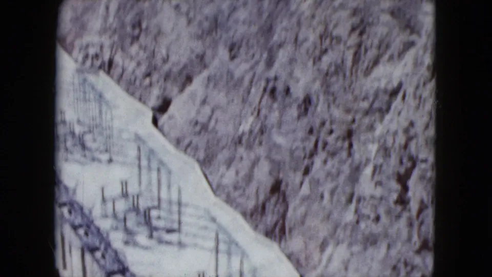 1959 Nevada 8mm frame of a rocky hillside, pale road or wash, and small vertical posts in dry terrain.