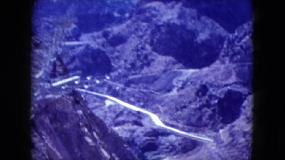 Elevated 1959 Nevada 8mm frame of canyon terrain, dark blue water, a pale pipeline or road, and rugged exposed slopes.