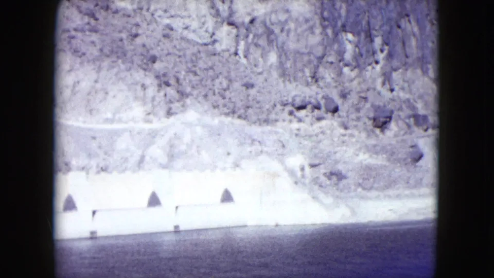 1959 Nevada 8mm view of a pale concrete dam or retaining structure beside dark water under a rocky canyon slope.