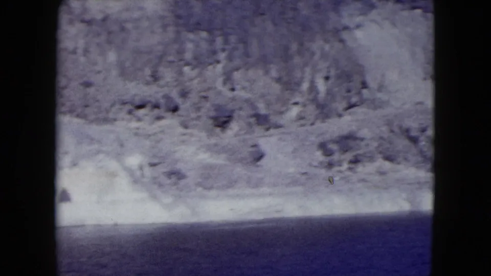 1959 Nevada 8mm frame showing pale rocky mountains dropping toward a deep blue lake or reservoir.