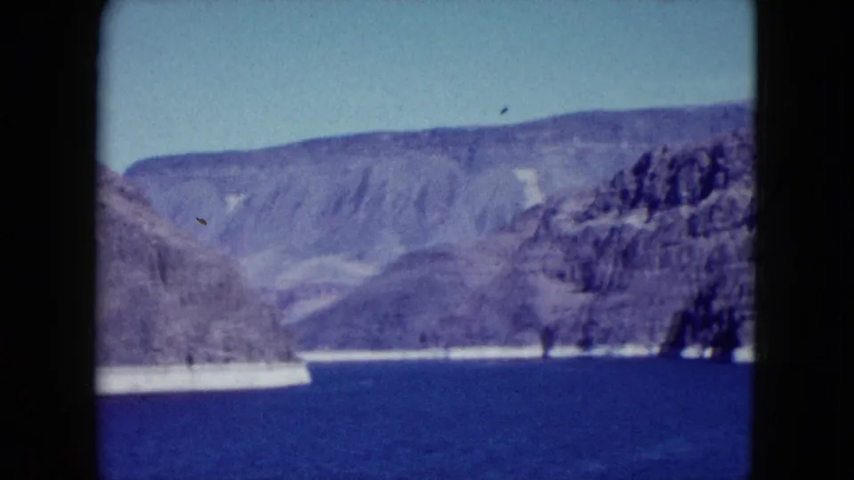 1959 Nevada home-movie frame showing a broad mountain-and-water vista with rocky slopes and bright shore.