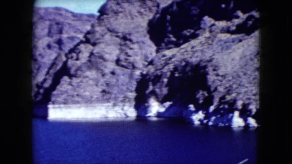 1959 Nevada 8mm frame showing a closer rocky shoreline above blue water, with purple-blue film color shift and soft detail.