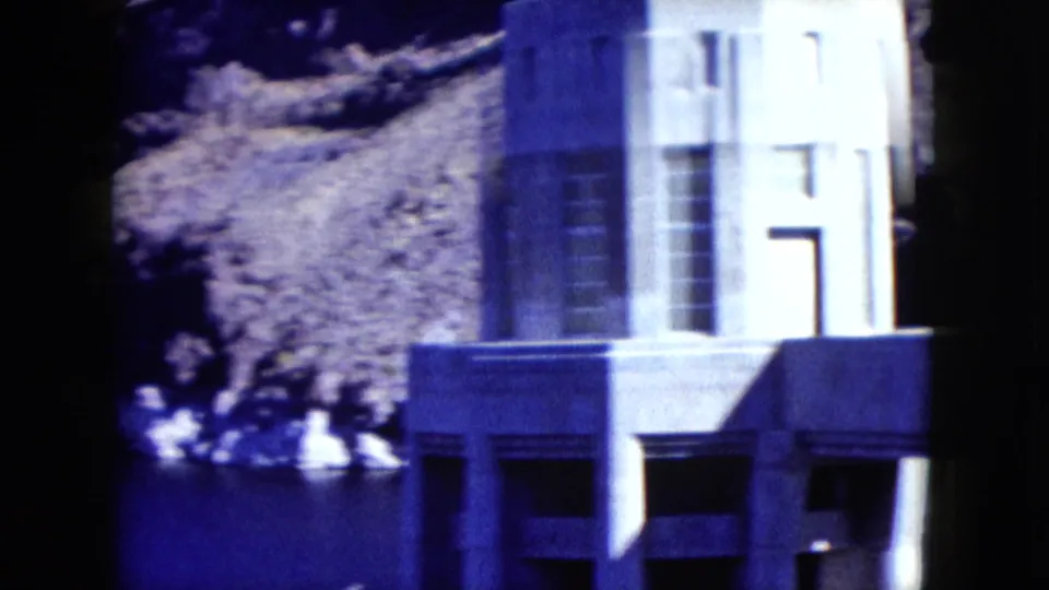 1959 Nevada home-movie frame with a white concrete dam or water-intake structure at the edge of blue water near rocky terrain.