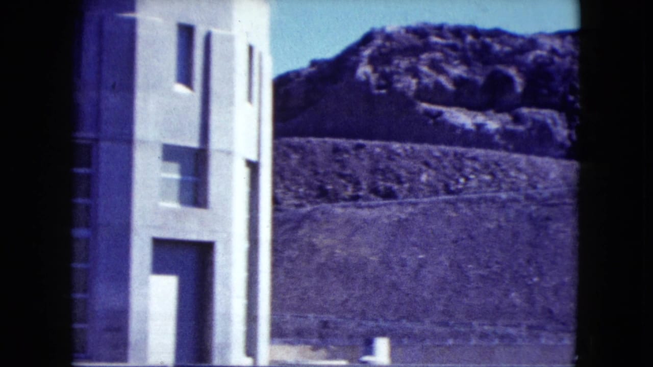 1959 Nevada 8mm frame showing a pale concrete industrial building near fencing with rugged desert hills in the background.