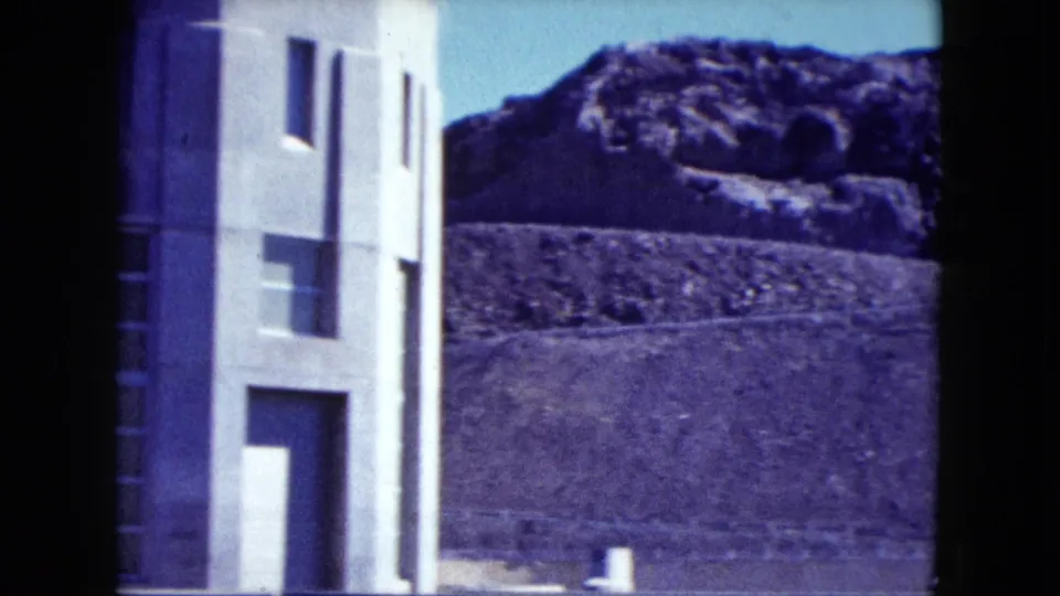 1959 Nevada 8mm frame showing a pale concrete industrial building near fencing with rugged desert hills in the background.