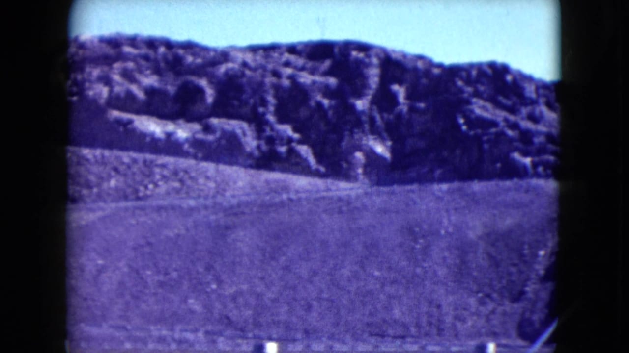 1959 Nevada 8mm view across a rocky desert hillside and open graded slope near an industrial site.