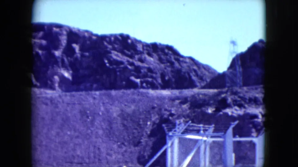 1959 Nevada 8mm frame showing fencing, utility structures, rocky desert hills, and a distant tower on the ridge.