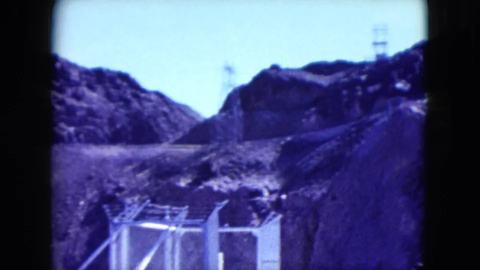 1959 Nevada 8mm view of fenced utility structures at the base of rugged desert hills with a tower visible on the skyline.