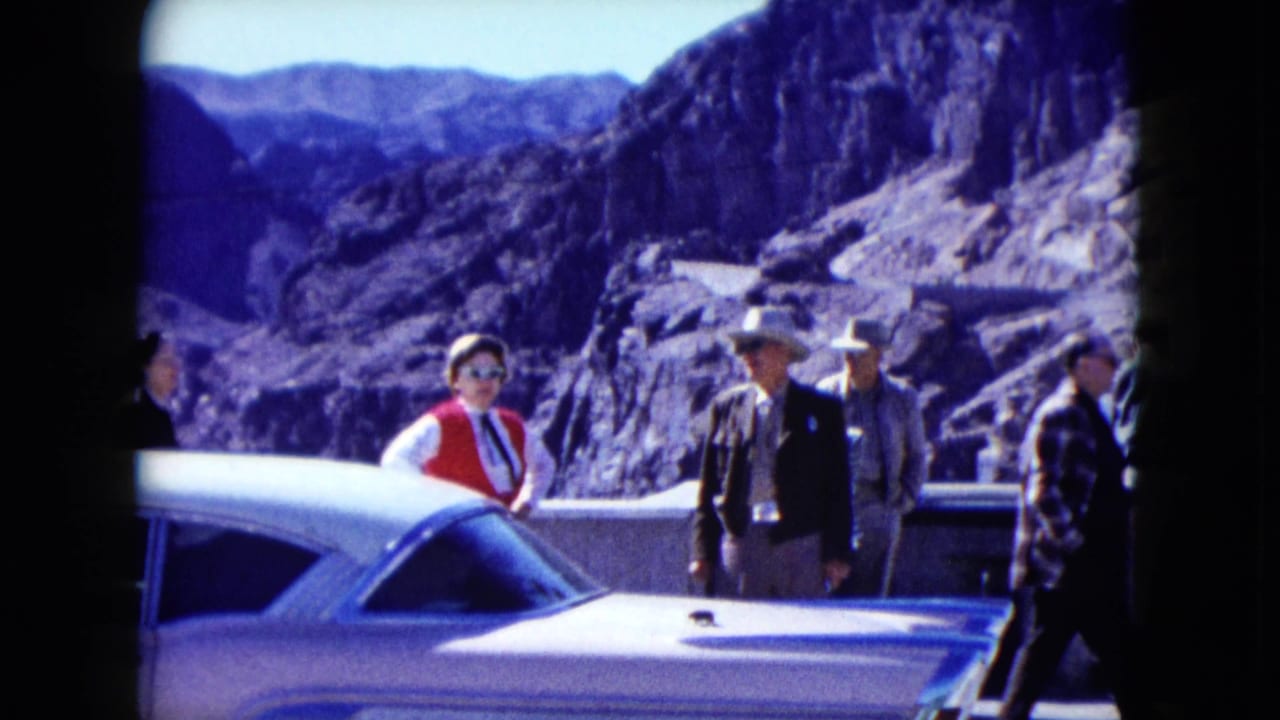 1959 Nevada home-movie frame showing tourists near a white classic car at a dam overlook with mountains behind them.
