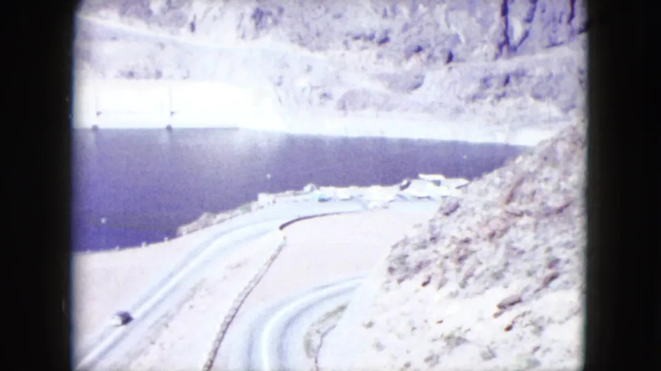 1959 Nevada home-movie view looking down toward a reservoir, dam structure, pale slopes, and winding access roads.