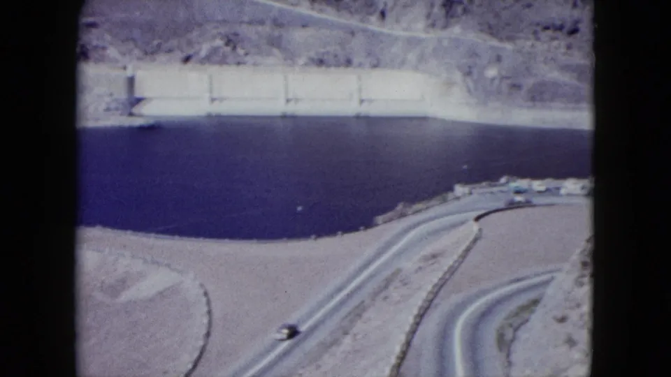 Wide 1959 Nevada view of dark reservoir water, a dam wall, pale rocky terrain, and winding roads from a high overlook.