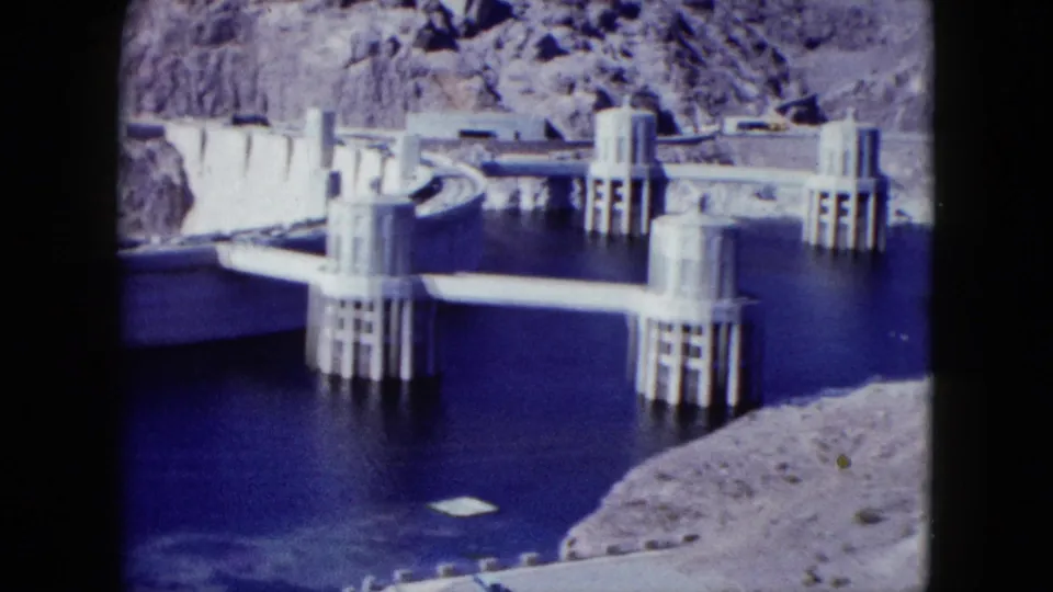 1959 Nevada 8mm frame centered on cylindrical intake towers, concrete spans, reservoir water, and desert shoreline.