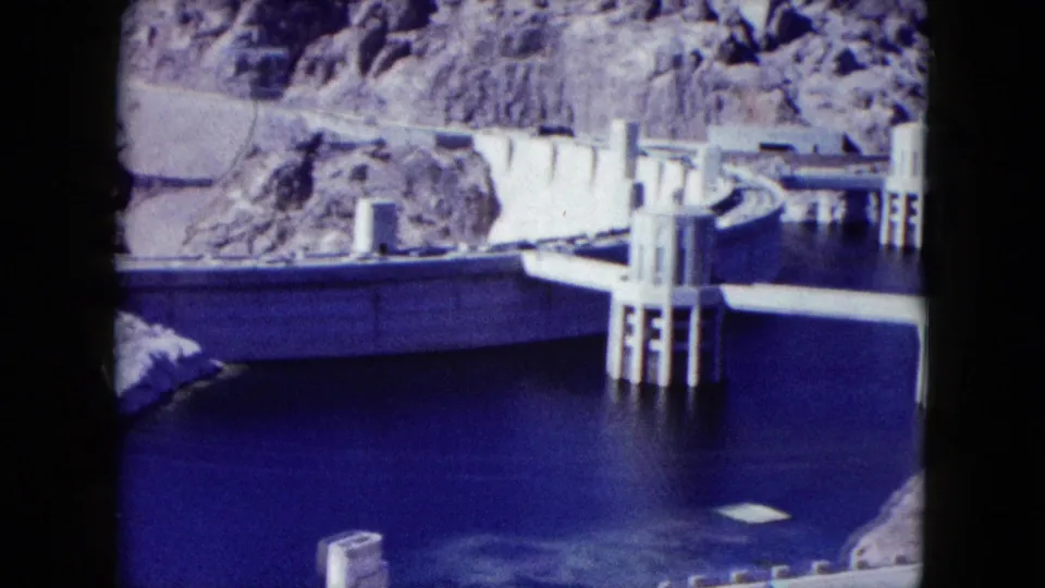 1959 Nevada 8mm view of a concrete dam wall, intake towers, deep blue reservoir water, and surrounding desert rock.