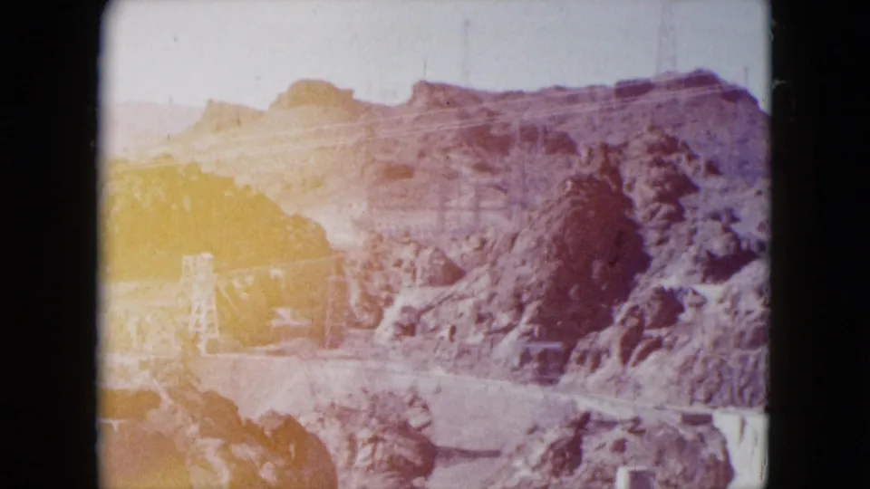 Faded 1959 Hoover Dam travel frame showing sunlit canyon rock, pale sky, and part of the dam structure below.