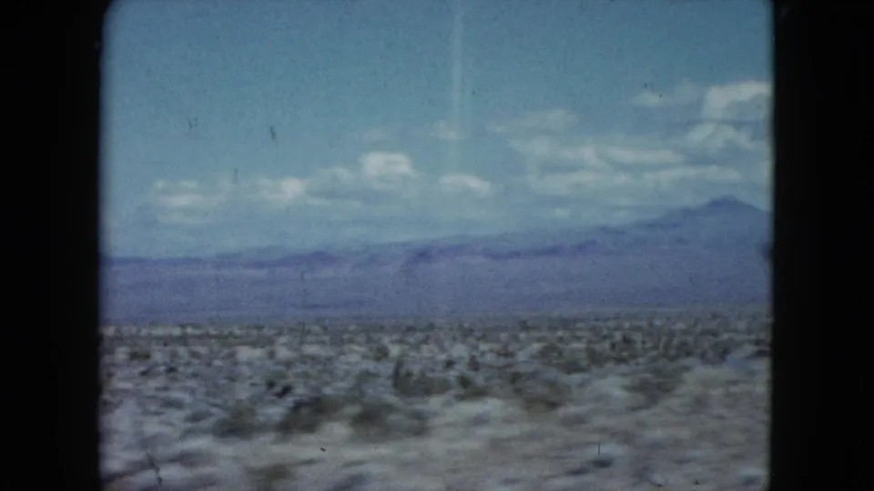 1959 Nevada 8mm view across low scrub desert toward hazy mountains under scattered clouds.