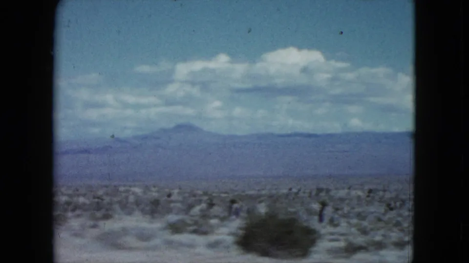1959 Nevada home-movie frame showing scrub-covered desert foreground and a hazy mountain ridge beyond.