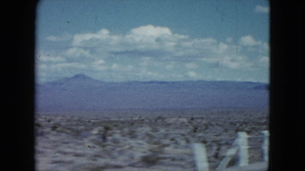 1959 Nevada desert roadside seen from a moving vehicle, with white fence posts, low scrub, distant mountains, and a blue sky.