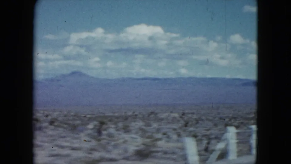 1959 Nevada desert roadside seen from a moving vehicle, with white fence posts, low scrub, distant mountains, and a blue sky.