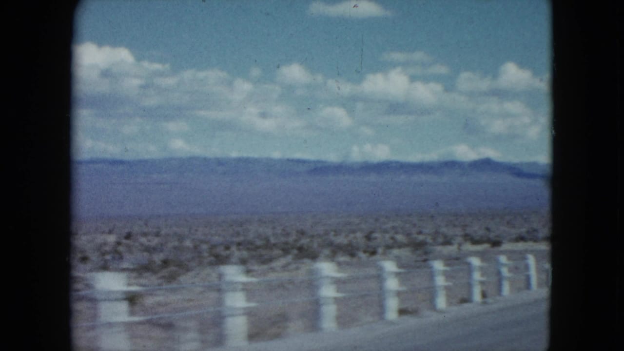 Passenger-view 8mm frame of the Nevada desert, showing a roadside fence, open scrubland, distant mountains, and clouded blue sky.