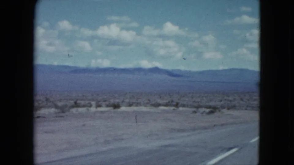 1959 Nevada highway view from a moving car, with pavement in the foreground, desert scrub, mountains, and scattered clouds.