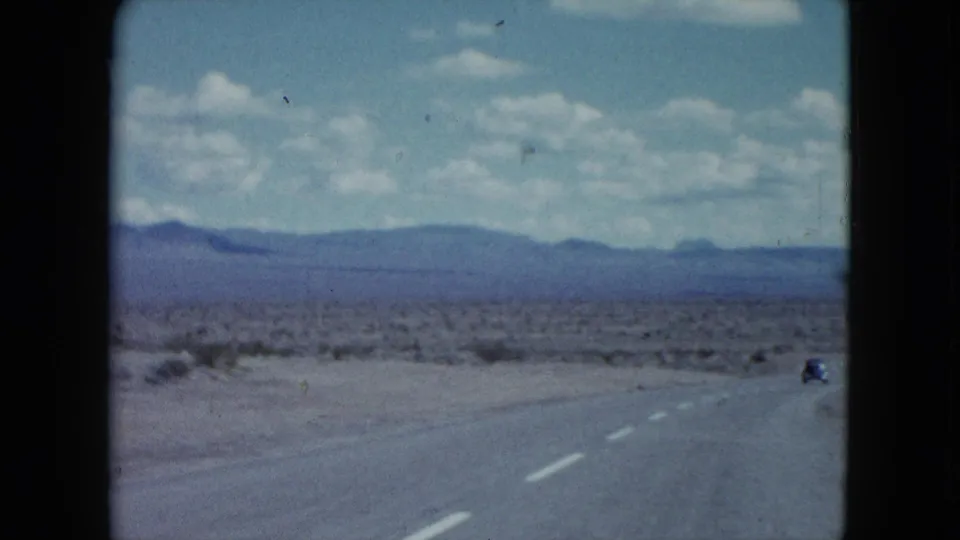 Nevada desert road in 1959 with a small distant car, lane markings, scrubland, mountains, and scattered clouds.