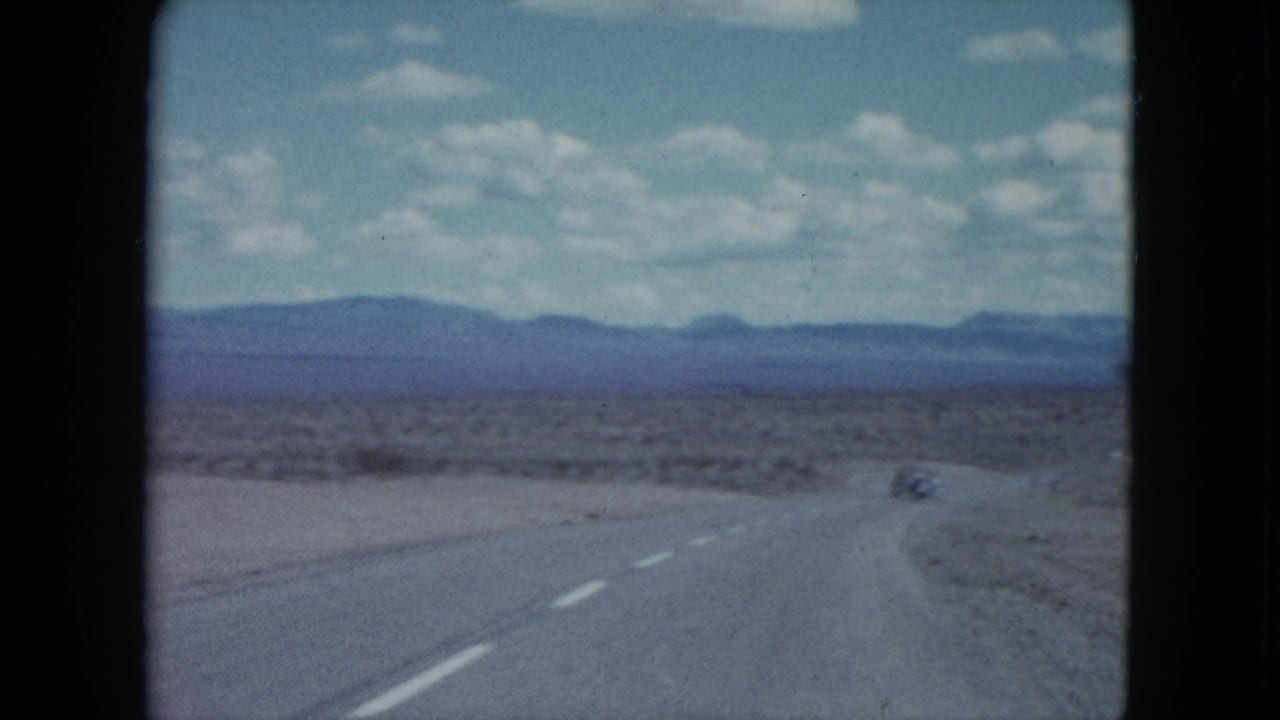 1959 Nevada home-movie frame showing a paved desert road, distant vehicle, low mountains, and scattered clouds.