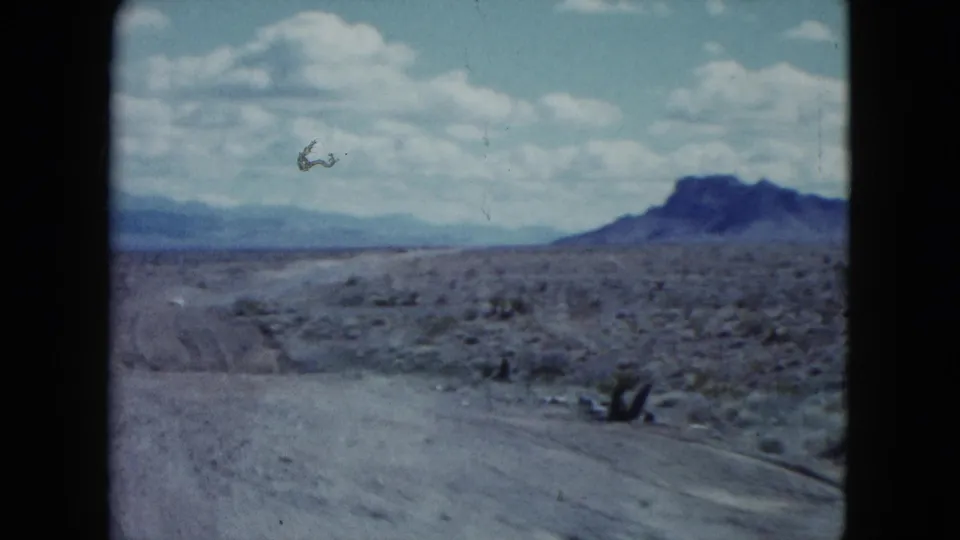 1959 Nevada desert view with a curving roadside, sparse vegetation, distant mountains, and a brief bird-like shape in the sky.