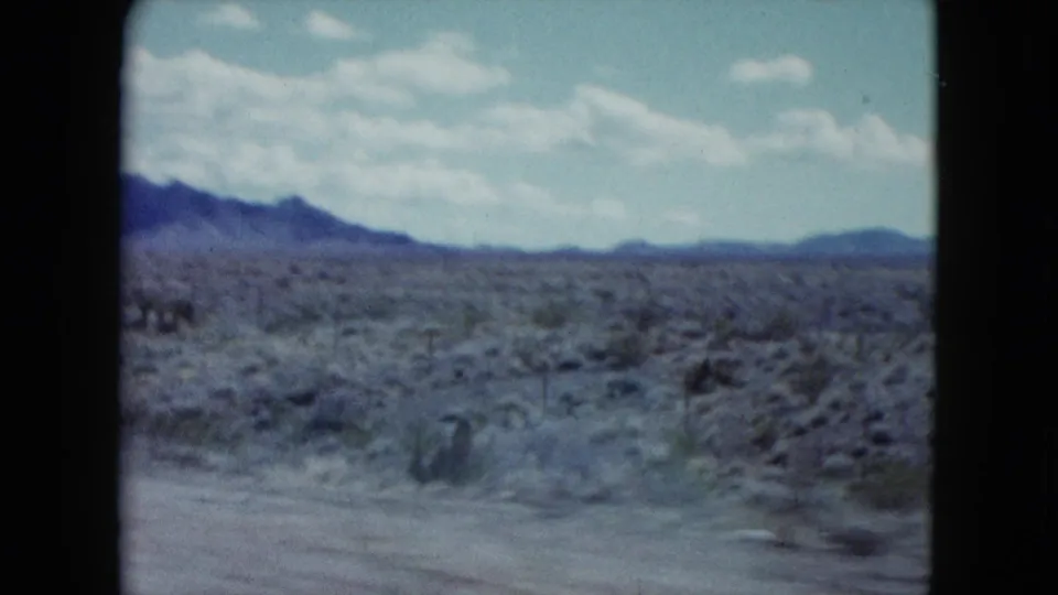 1959 Nevada home-movie frame showing open desert scrubland, a roadside edge, low mountains, and a bright clouded sky.