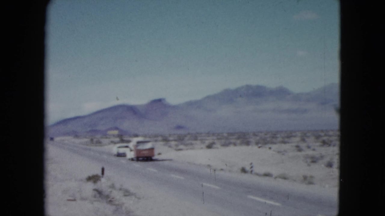 A car towing a red-and-white camper trailer travels along a desert highway with mountains in the distance.