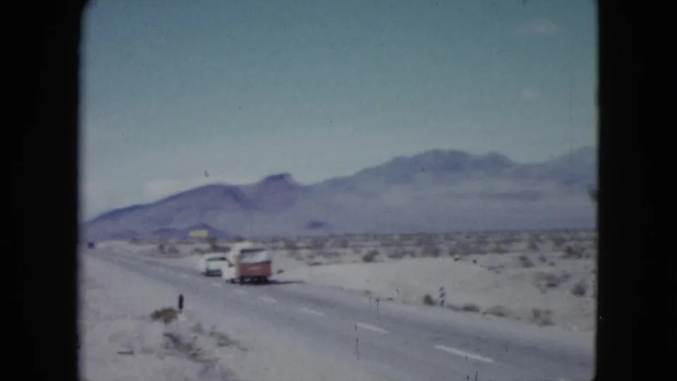 1959 Nevada home-movie frame showing a white car pulling a red-and-white camper trailer on a two-lane desert road below distant mountains.