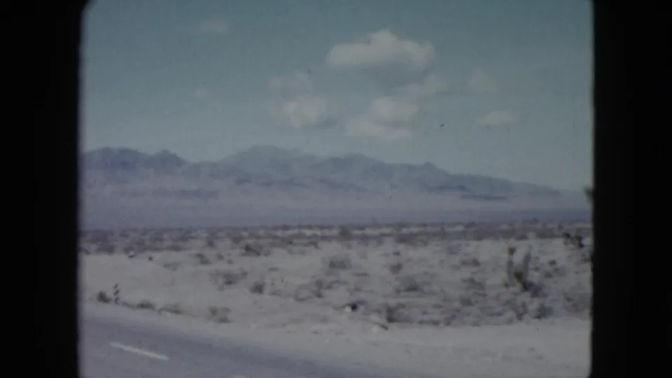 1959 Nevada desert landscape frame with roadside pavement, low scrub, hazy mountains, and soft color from an 8mm home movie scan.