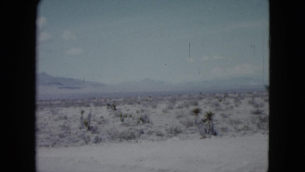 1959 Nevada 8mm frame showing a whitish desert plain, sparse shrubs, and hazy mountains beneath a pale blue sky.