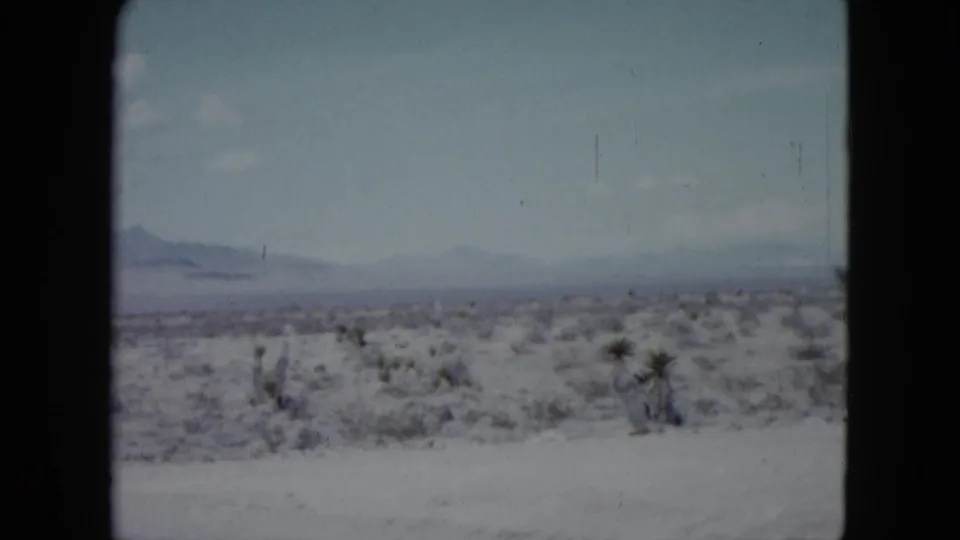 1959 Nevada 8mm frame showing a whitish desert plain, sparse shrubs, and hazy mountains beneath a pale blue sky.