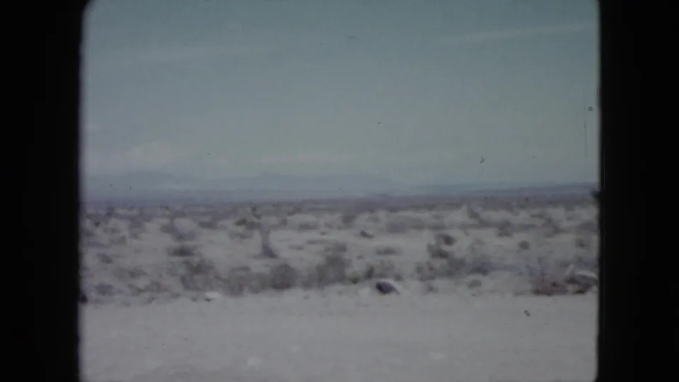 1959 Nevada home-movie frame with faded color, sparse brush, a flat pale desert floor, and distant mountains under thin cloud.