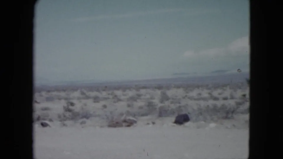 Nevada desert landscape on 8mm film, with scrub plants, scattered dark rocks, whitish ground, and mountains fading into the horizon.