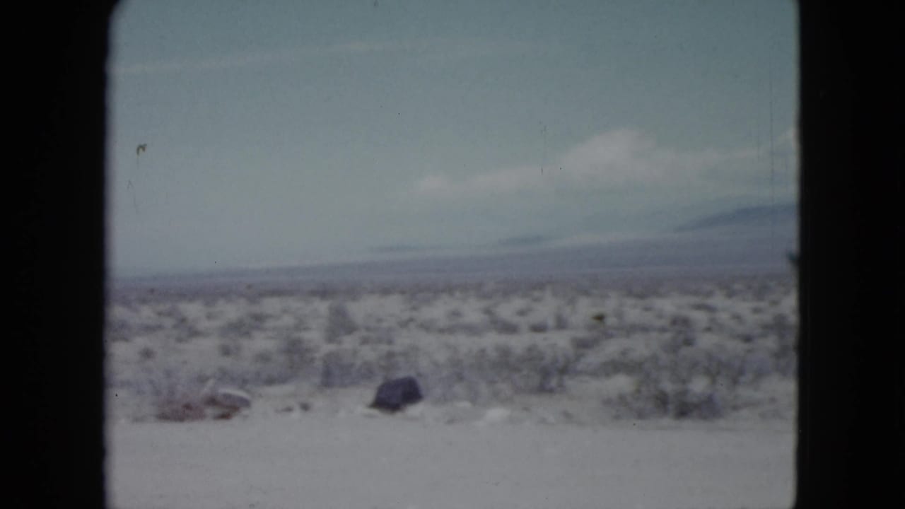 1959 Nevada 8mm frame showing open desert scrub, pale ground, scattered rocks, and soft distant mountains beneath light cloud.