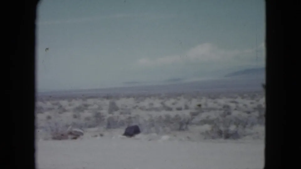 1959 Nevada 8mm frame showing open desert scrub, pale ground, scattered rocks, and soft distant mountains beneath light cloud.
